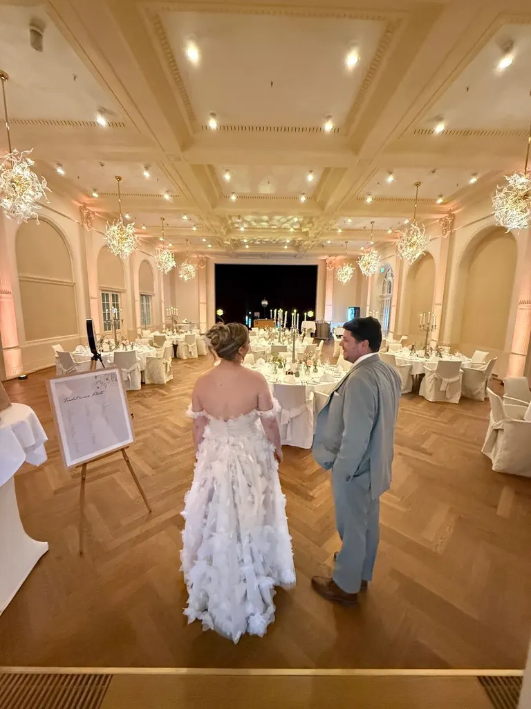 Brautpaar steht im Festsaal und blickt über die gedeckten Tische, links eine Sitzplan-Tafel