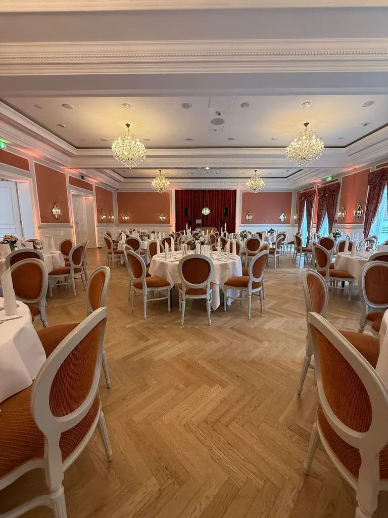 Festsaal mit gedeckten runden Tischen, Polsterstühlen und Kronleuchtern unter der Stuckdecke