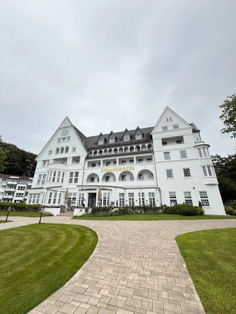 Weiße Fassade des Strandhotels Glücksburg mit gepflastertem Weg und Rasen unter bewölktem Himmel